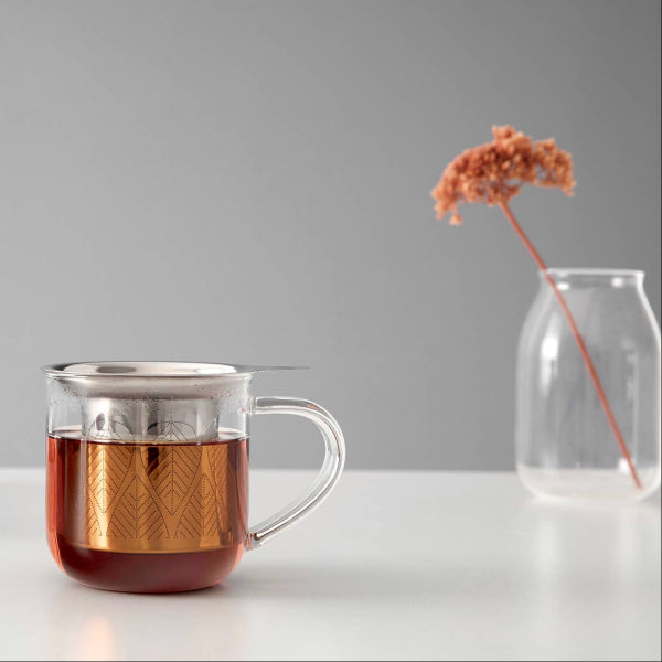 Tea cup with a  stainless steel infuser, and vase on a white surface with gray background. The Spice & Tea Shoppe