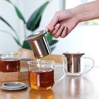 Person preparing tea in a glass mug with a stainless steel infuser on a wooden tray. The Spice & Tea Shoppe