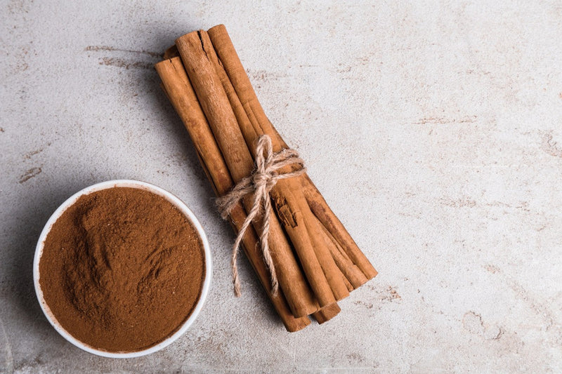 Ground cinnamon in a white bowl next to bundled cinnamon sticks on a light stone surface.