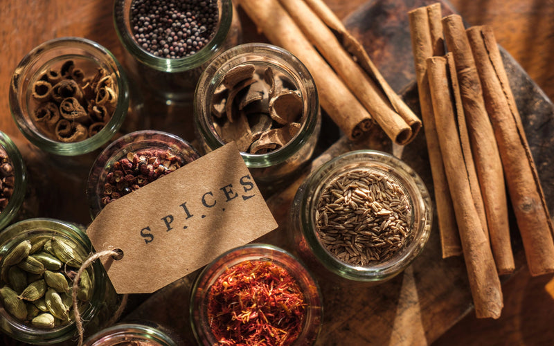 A top-down view of small glass jars filled with various whole spices, alongside cinnamon sticks and a kraft paper label reading "SPICES."