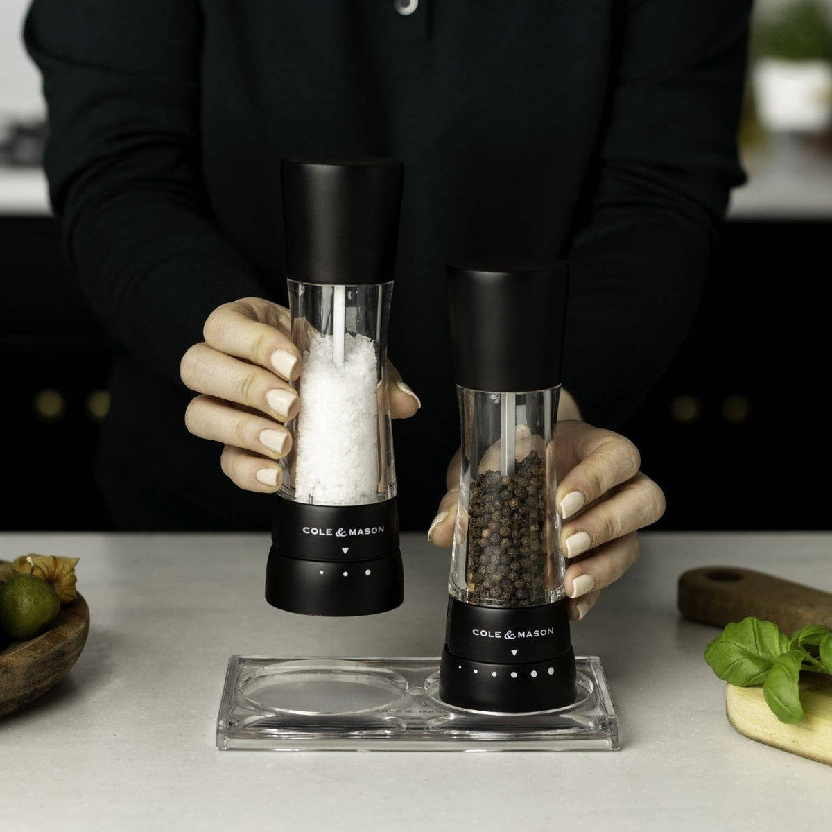 A person holding a pair of matte black Cole & Mason salt and pepper mills over a clear acrylic rest tray on a kitchen counter.