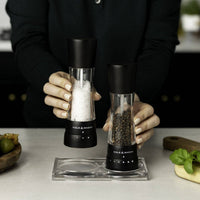 A person holding a pair of matte black Cole & Mason salt and pepper mills over a clear acrylic rest tray on a kitchen counter.