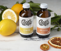 Two bottles of Cooks pure extract with lemons and oranges on a marble surface.