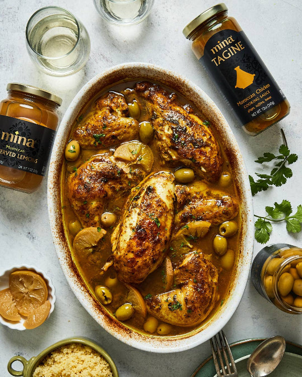 Dish of chicken tagine with olives and lemon, surrounded by jars of mina Tagine and spices on a light background. The Spice & Tea Shoppe