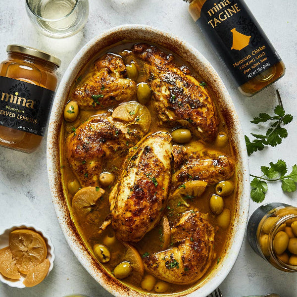 Dish of chicken tagine with olives and lemon, surrounded by jars of mina Tagine and spices on a light background.  The Spice & Tea Shoppe
