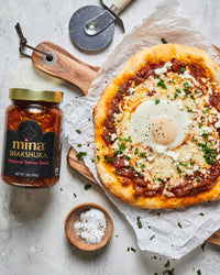 Pizza with Shakshuka topping next to a jar of Mina Shakshuka tomato sauce on a light surface. The Spice & Tea Shoppe