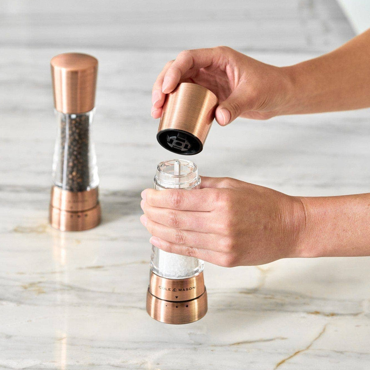A pair of Cole & Mason Derwent salt and pepper mills in a metallic copper finish being filled.