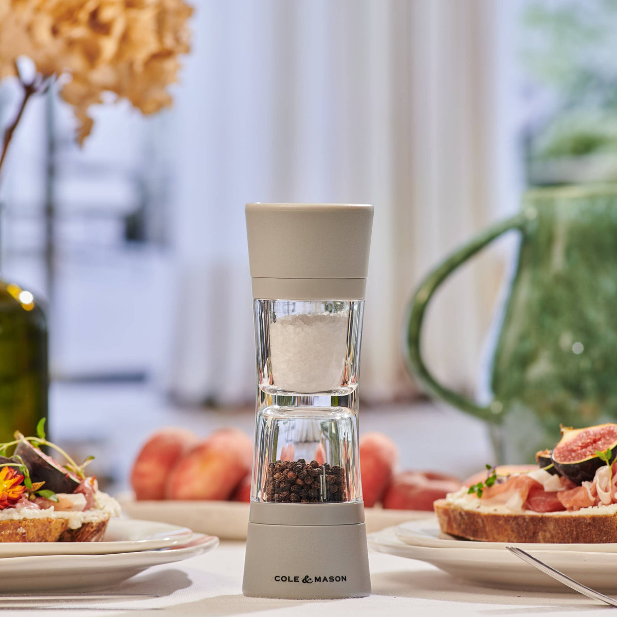A Cole & Mason Lincoln Duo salt and pepper mill standing vertically on a dining table surrounded by plates of food.