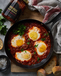 Shakshuka with eggs in a red sauce in a skillet on a wooden board, with a bottle of 'mina Shakshuka' seasoning. The Spice & Tea Shoppe