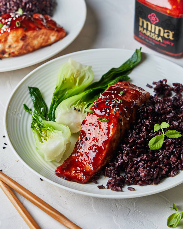 Plate of glazed salmon with black rice and greens, next to a bottle of Mina Harissa sauce. The Spice & Tea Shoppe