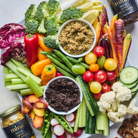 Platter of fresh vegetables with two jars of dip on a light gray background