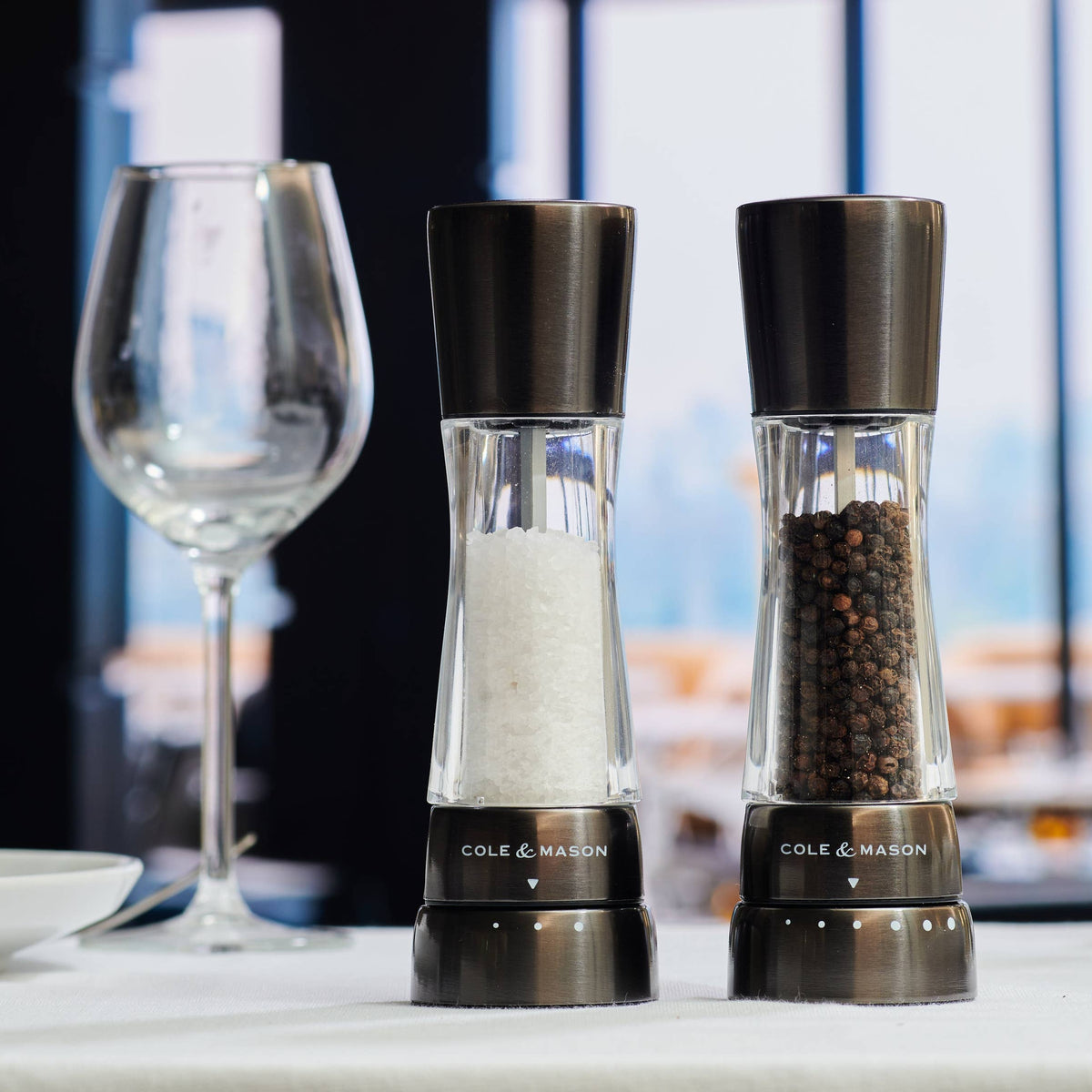A pair of Cole & Mason Derwent salt and pepper mills in a sleek, gunmetal grey finish illuminated by a kitchen window on a white tablecloth with a wine glass in the background.