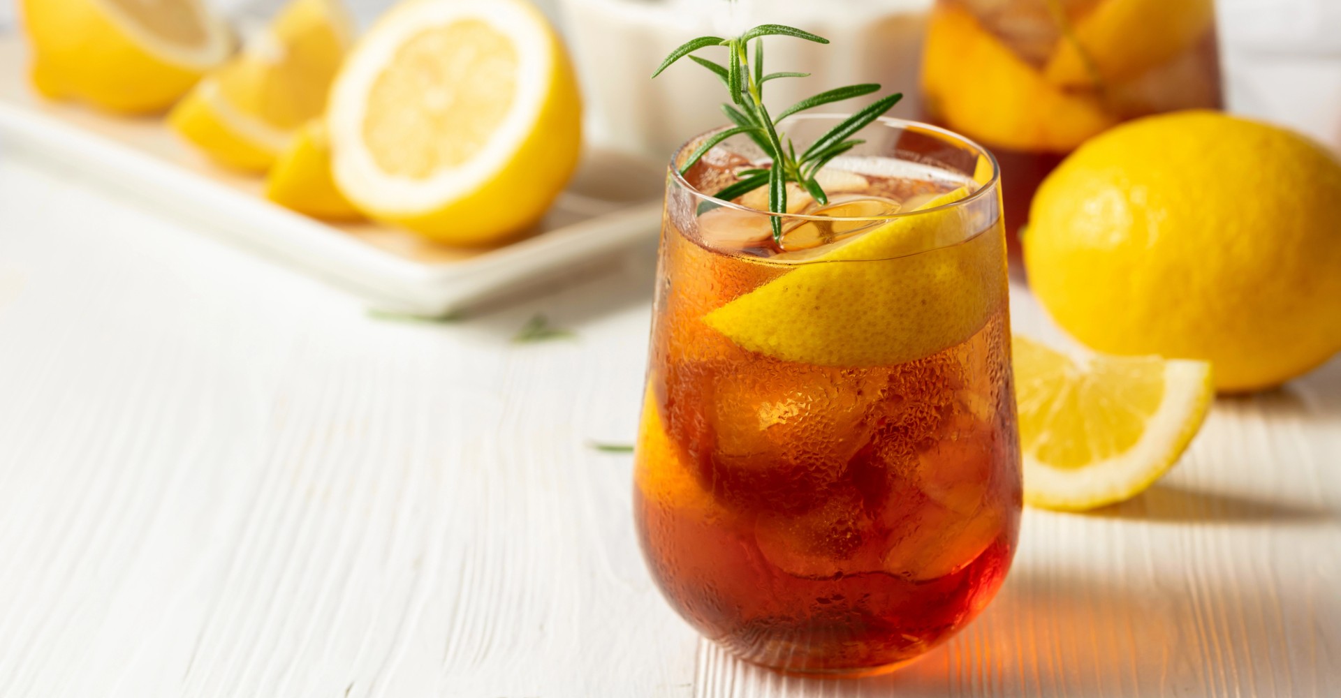 Glass of iced tea with lemon slices and a sprig of rosemary on a white surface with lemons in the background. A jar of Castellino Grilled Olives, with the brand name and product title visible on the label. The Spice & Tea Shoppe