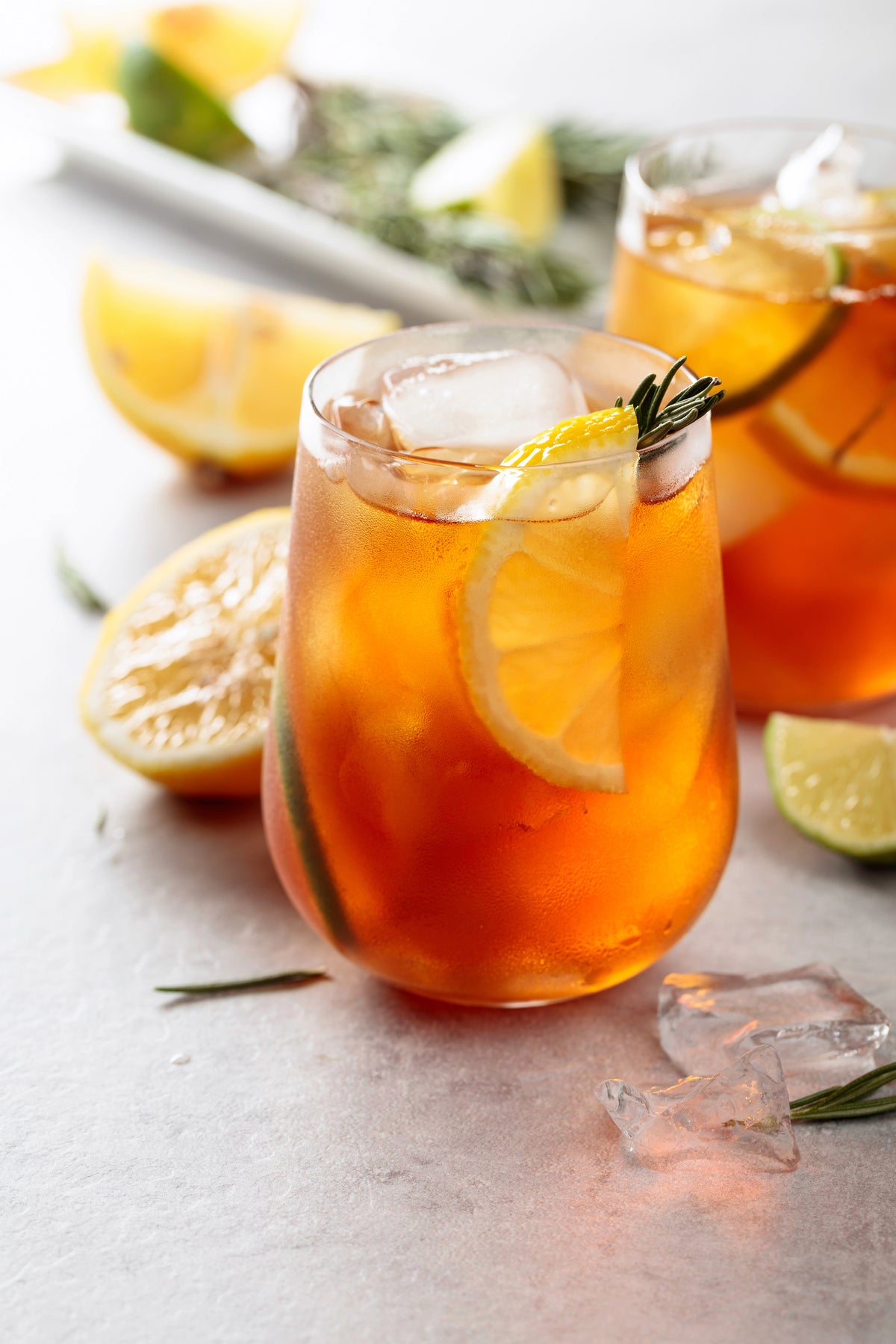 Glass of iced tea with lemon and ice cubes on a light surface with lemons and limes. A jar of Castellino Grilled Olives, with the brand name and product title visible on the label. The Spice & Tea Shoppe