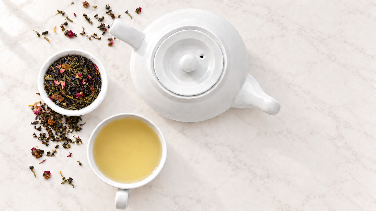 White teapot, cup of green tea, and loose tea leaves on warm ivory marble with soft natural light.