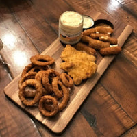 Assorted fried snacks on a wooden board with a jar of dipping sauce on a wooden table.