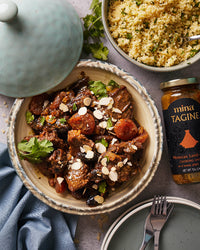 Bowl of lamb tagine with apricots and almonds, jar of mina tagine seasoning, and bowl of couscous on a gray surface.  The Spice & Tea Shoppe