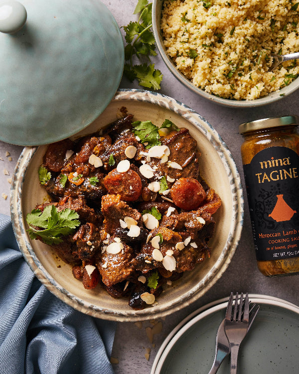 Bowl of lamb tagine with apricots and almonds, jar of mina tagine seasoning, and bowl of couscous on a gray surface.  The Spice & Tea Shoppe