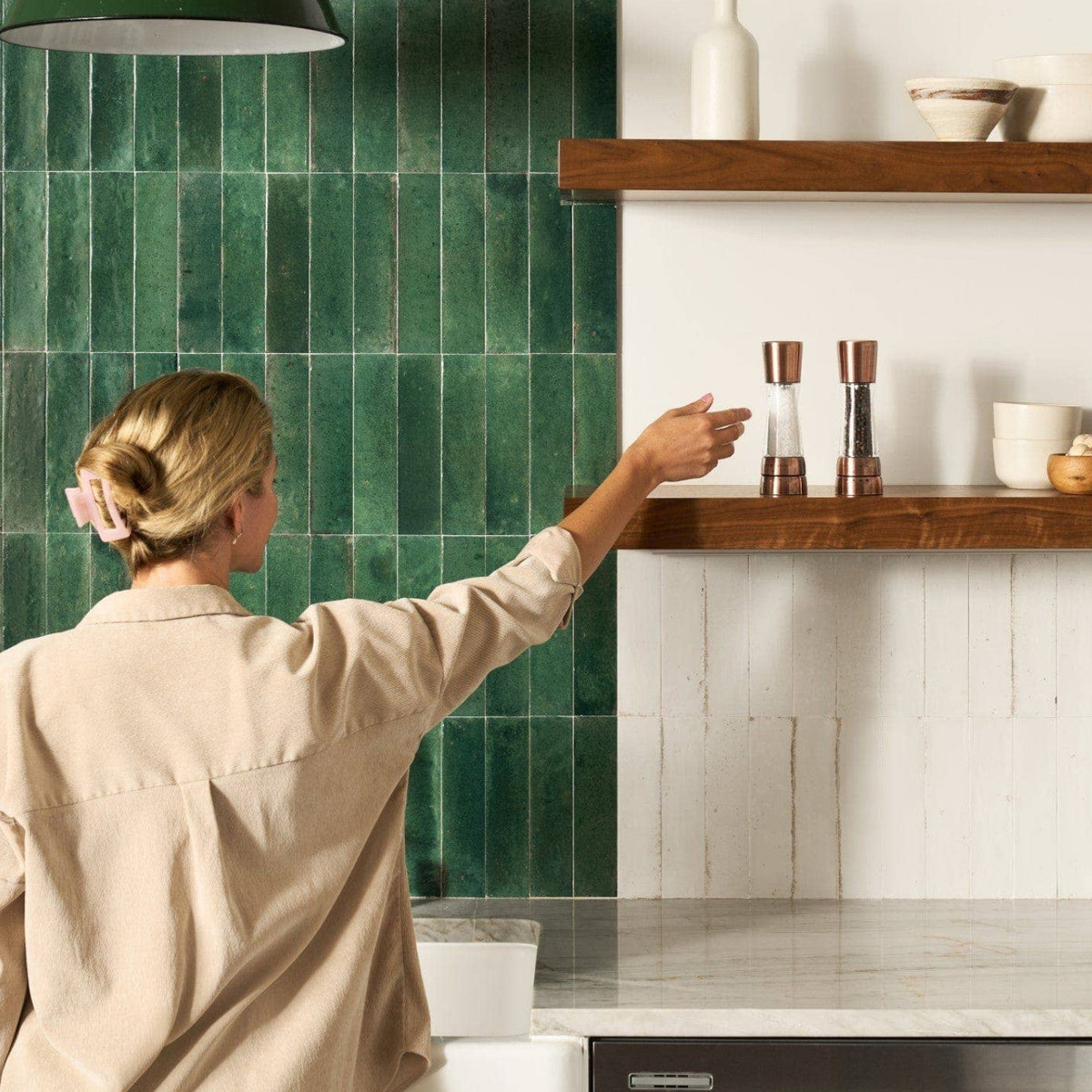 A woman reaching toward a pair of copper Cole & Mason salt and pepper mills on a wooden floating shelf in a modern kitchen.

