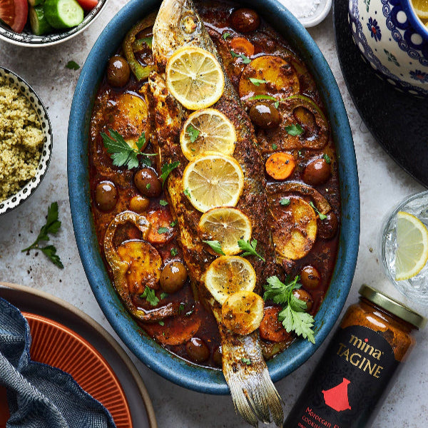 Baked fish dish with lemon slices and olives in a blue oval dish on a table with side dishes. The Spice & Tea Shoppe