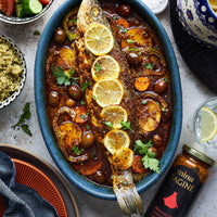 Baked fish dish with lemon slices and olives in a blue oval dish on a table with side dishes. The Spice & Tea Shoppe