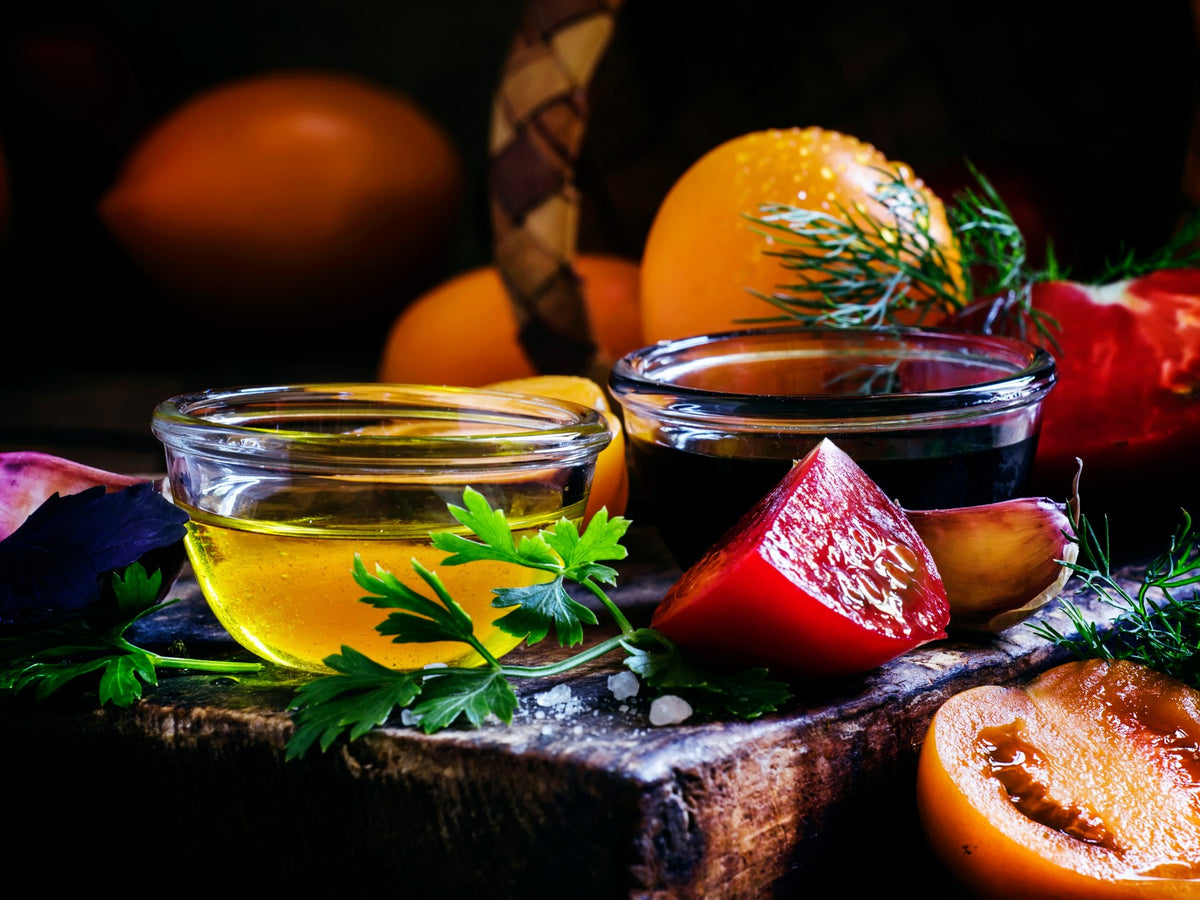 Small glass bowls of golden olive oil and dark balsamic vinegar sit on a rustic wooden board, surrounded by fresh herbs, sea salt, a halved red tomato, sliced melon, and whole stone fruits in the background. The composition highlights vibrant colors and fresh ingredients. The Spice & Tea Shoppe